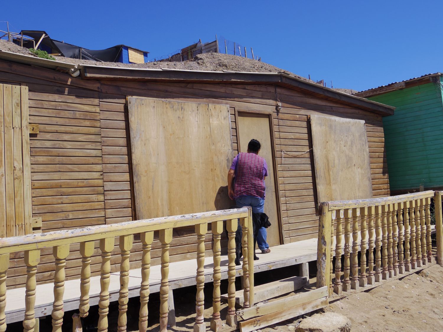 砂浜に立つ手すり付きの簡素なベランダがある木造の家の扉の前に、女性が黒い犬とともに立っている写真