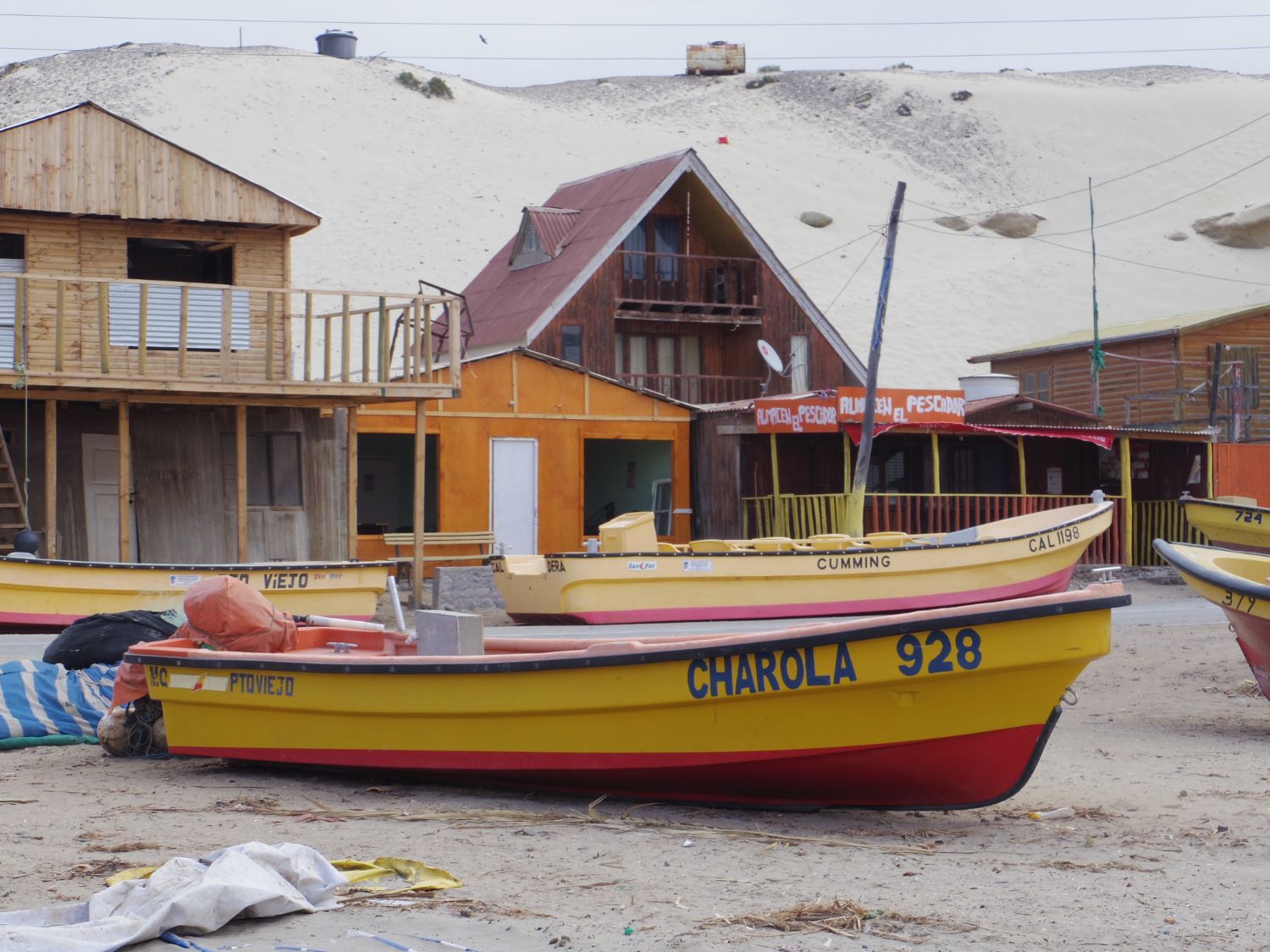 「CHAROLA 928」と書かれた黄色と赤の漁船が砂浜にあり、背後にはカラフルな木造の家々や砂丘が見える写真