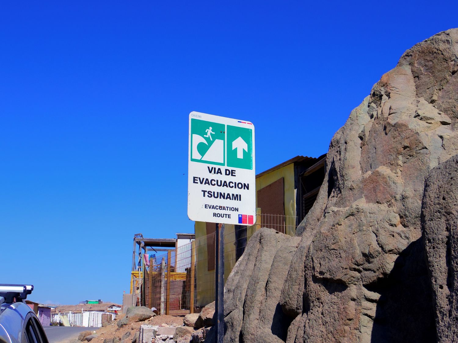 青空の下に『VIA DE EVACUACION TSUNAMI(津波避難路)』と書かれた標識が立ち、その背後には岩場と住宅が見える、津波時の避難経路を示す案内標識の写真