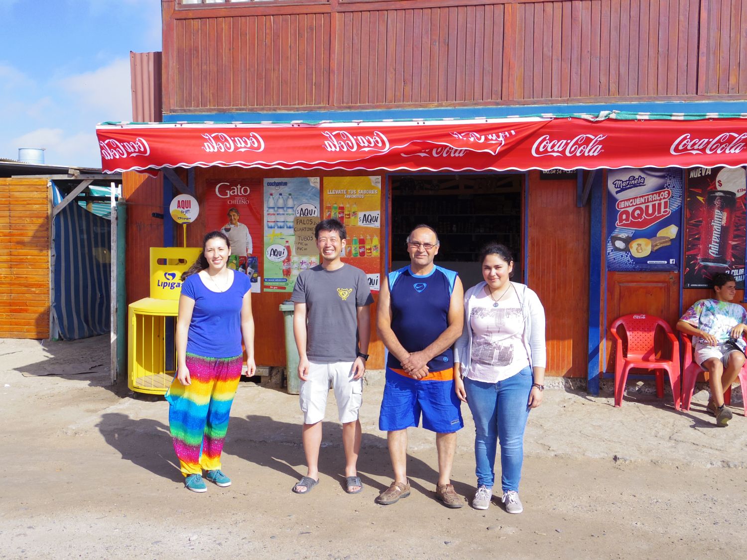 木造の店の前で4人の男女が並んで笑顔で立ち、看板にはコカコーラやアイスの広告が並ぶお店の写真