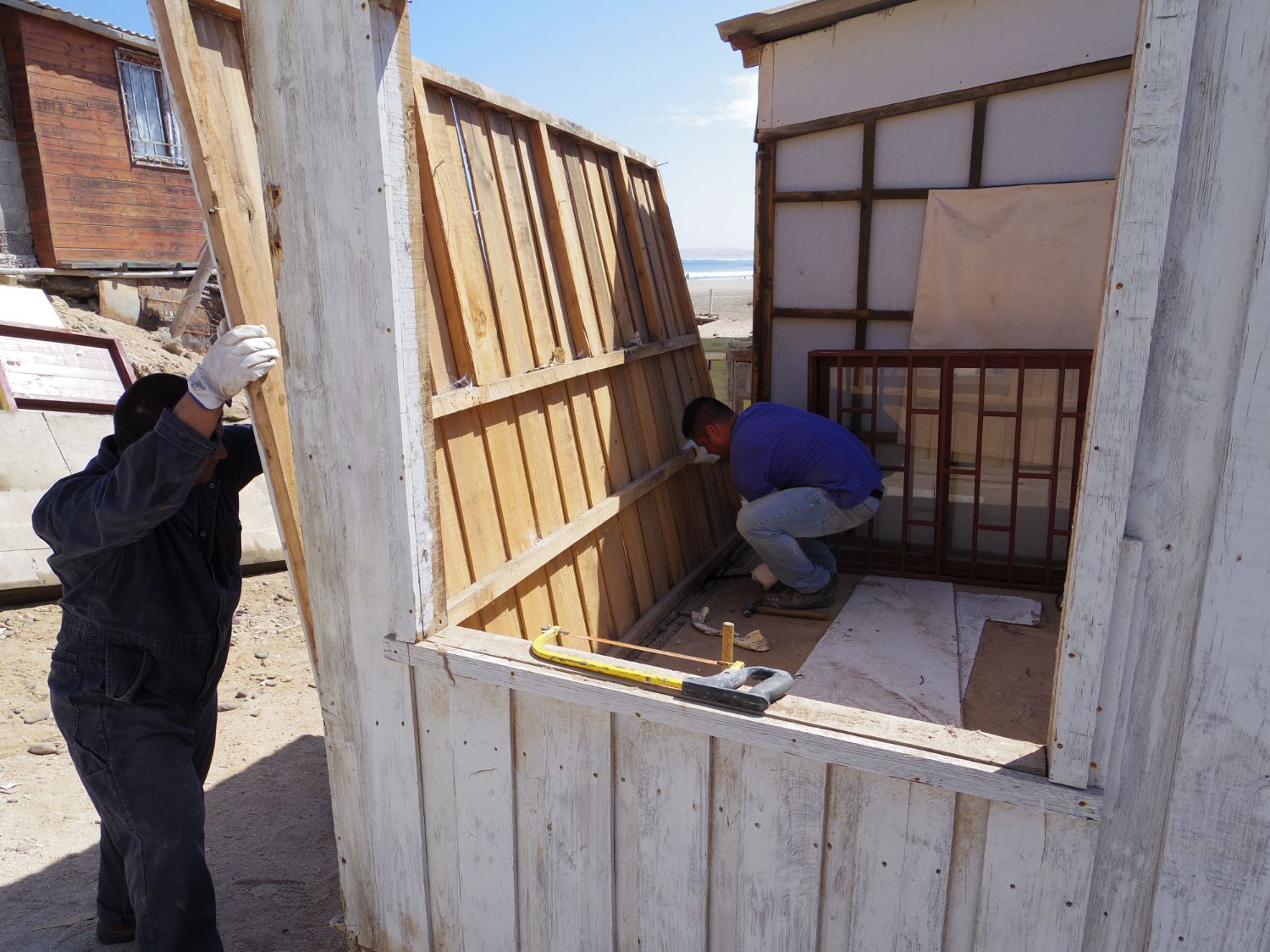 砂地の中で日差しを浴びながら作業する二人の男性が木造の小屋を組み立てている様子の写真