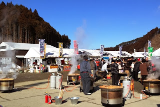 屋外のイベント会場で大きな釜から蒸気が立ち上り、多くの人が牡蠣料理を楽しんでいる様子の写真