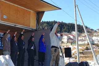 ステージの上でマイクを持った男性と関係者たちが拳を掲げてイベントの開会を宣言している様子の写真