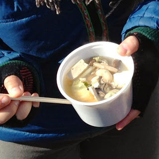 牡蠣や豆腐、野菜などの入った温かいスープの入った器を持っている手元を写した写真