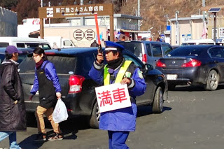 駐車場の出入口で警備員が「満車」と書かれた札を掲げて案内し、後方には車と人の往来がある様子の写真