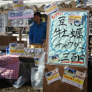 テントの前で「名物 牡蠣チャウダー」と値段が書かれた看板とともに店舗の男性が写っている写真