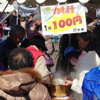 屋外のイベント会場で「カキ汁1杯100円」と書かれた看板の下、スタッフが大鍋から湯気の立つカキ汁を来場者に盛りつけて提供している様子の写真