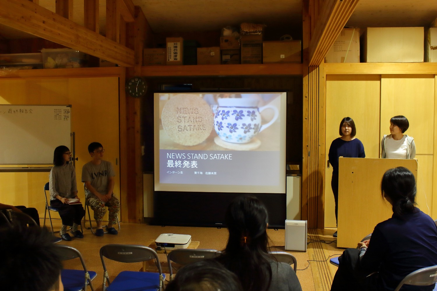 木造の温かみのある室内で、スクリーンに「NEWS STAND SATAKE 最終発表」と表示される中、2人の女性が前に立って発表し、他の参加者が椅子に座ってそれを真剣に見守っている様子のプレゼンテーションの写真
