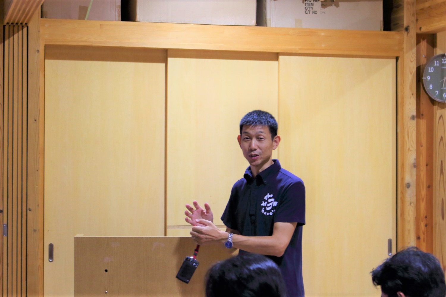 木の内装に囲まれた室内で、黒いポロシャツを着た男性が演台の前に立ち、手振りを交えて聴衆に語りかけるように発表している写真