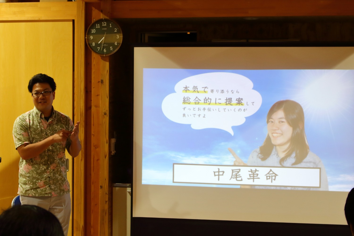 木の温もりを感じる室内で、男性が笑顔でプレゼンを行い、スクリーンには女性の写真と「本気で寄り添うなら総合的に提案してずっとお手伝いしていくのが良いですよ」という吹き出しと「中尾革命」の文字が大きく映し出されている写真