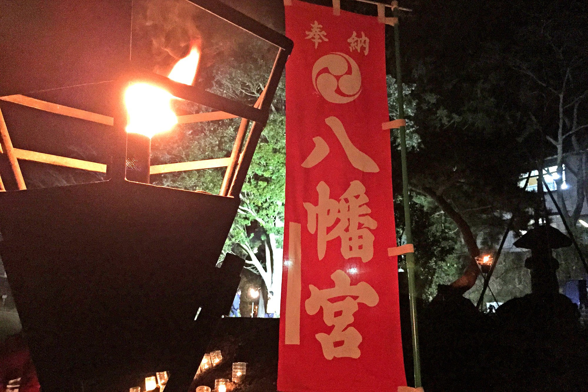 夜の屋外で、左側に大きなかがり火が燃えているのが明るく写り、その隣に赤い布に白い文字で「八幡宮」と書かれた旗が立っている写真