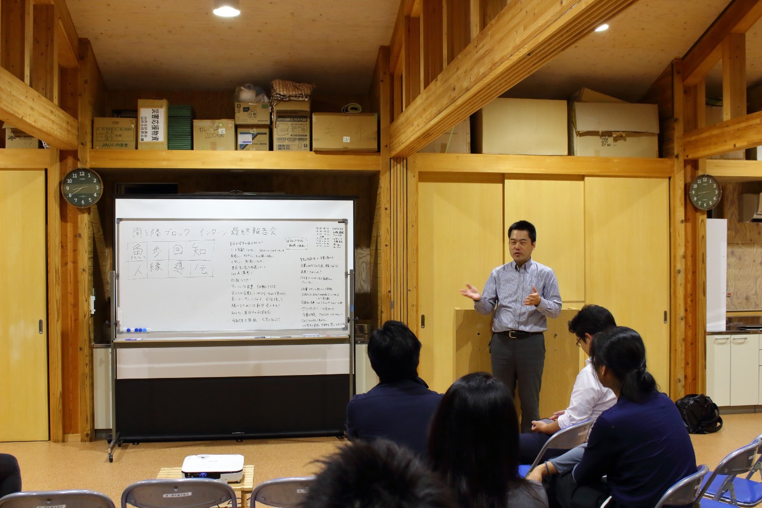 木の温もりを感じる会議室で、ホワイトボードに「南三陸ブロック インターン最終報告会」と書かれ、左側にはキーワードがマス目状に、右側には多くのメモや発言が書かれている前で、スーツ姿の男性が参加者に向けて身振りを交えて真剣に話している写真