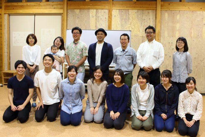 15人の大人と1人の赤ちゃんが、木の壁と床のある室内でリラックスした雰囲気で笑顔を見せながらポーズをとっている集合写真