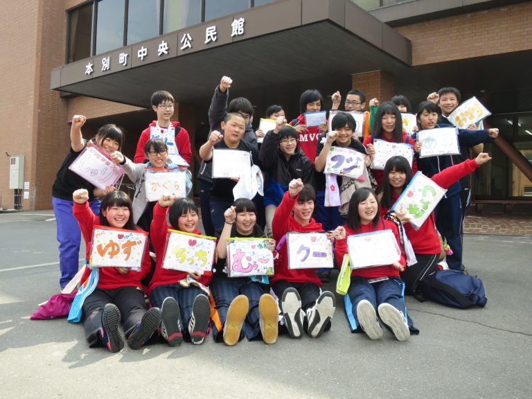 「本別町中央公民館」と書かれた茶色の外壁の建物入り口の前で、ニックネームのような名前が書かれた透明ケースをそれぞれ持った生徒たちが、拳を握った右手を上げている集合写真