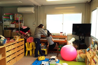 棚やおもちゃが並んだ室内で、大人の女性がピアノに向かって座る子どもに手を添えて弾き方を教えている写真