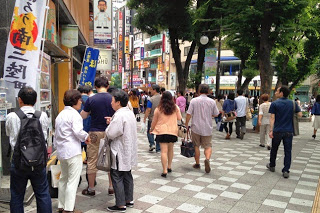 商店街の歩道を多くの人が行き交い、「宮城ふるさとプラザ」の前で立ち止まって商品を見ている人々の様子の写真