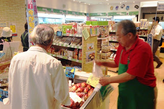 緑色のエプロンを着用した男性スタッフが冷蔵ケースの上でホヤのような商品を販売し、客に説明しながら渡している様子の写真