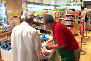 白いシャツを着た買い物客の男性に対し、赤い服と緑のエプロンの男性店員がホヤの入った冷蔵ケースを前に商品説明をしている様子の写真