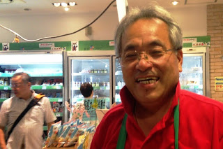 冷蔵棚の前の売り場に立ち、赤いシャツと緑のエプロンを身につけて笑顔で接客している男性の写真