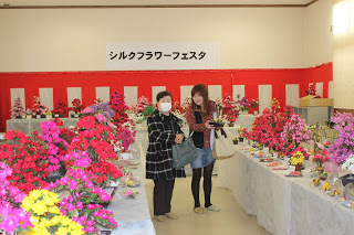 紅白幕が貼られた壁の上に「シルクフラワーフェスタ」と書かれた横断幕が掲げられた室内に、赤やピンク、黄色などの色とりどりのまゆ細工の花の作品が展示された白いクロスをかけた長机が並べられ、その間に、気に入った作品を手にした女性ら2人が立っている写真