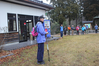 白い壁の建物の前にノルディックウォーキングポールを持ち、ピンク色のリュックサックを背負った男性が立ち、集まった人たちに話をしている様子の写真