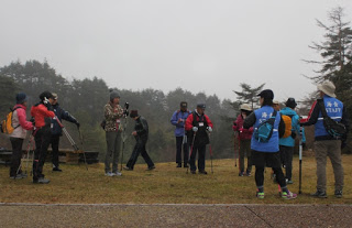 霧雨の中、森の前の広場にノルディックウォーキングポールを持った人々がたくさん集まっている写真