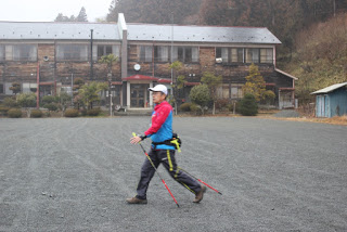 2階建ての建物前にある砂利の広場で、インストラクターの男性がノルディックウォーキングの歩き方の見本を見せている様子の写真