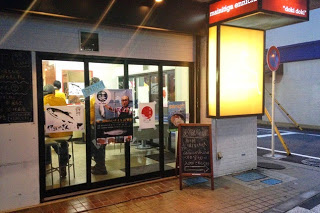 ガラス張りの店舗の外観で、中ではイベントが行われており、入口には黒板とポスターが貼られている、イベント中の飲食店の外観の写真