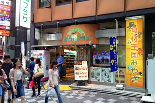 壁がオレンジ色のビルの一階にある「宮城ふるさとプラザ」の入口に人々が出入りし、右側には「食材王国みやぎ」と書かれた大きな看板が設置されている写真