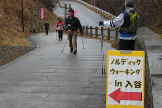 「ノルディックウォーキング in 入谷」の下に左向きの矢印が書かれた黄色い看板に向かって、参加者たちが坂を上ってきている写真