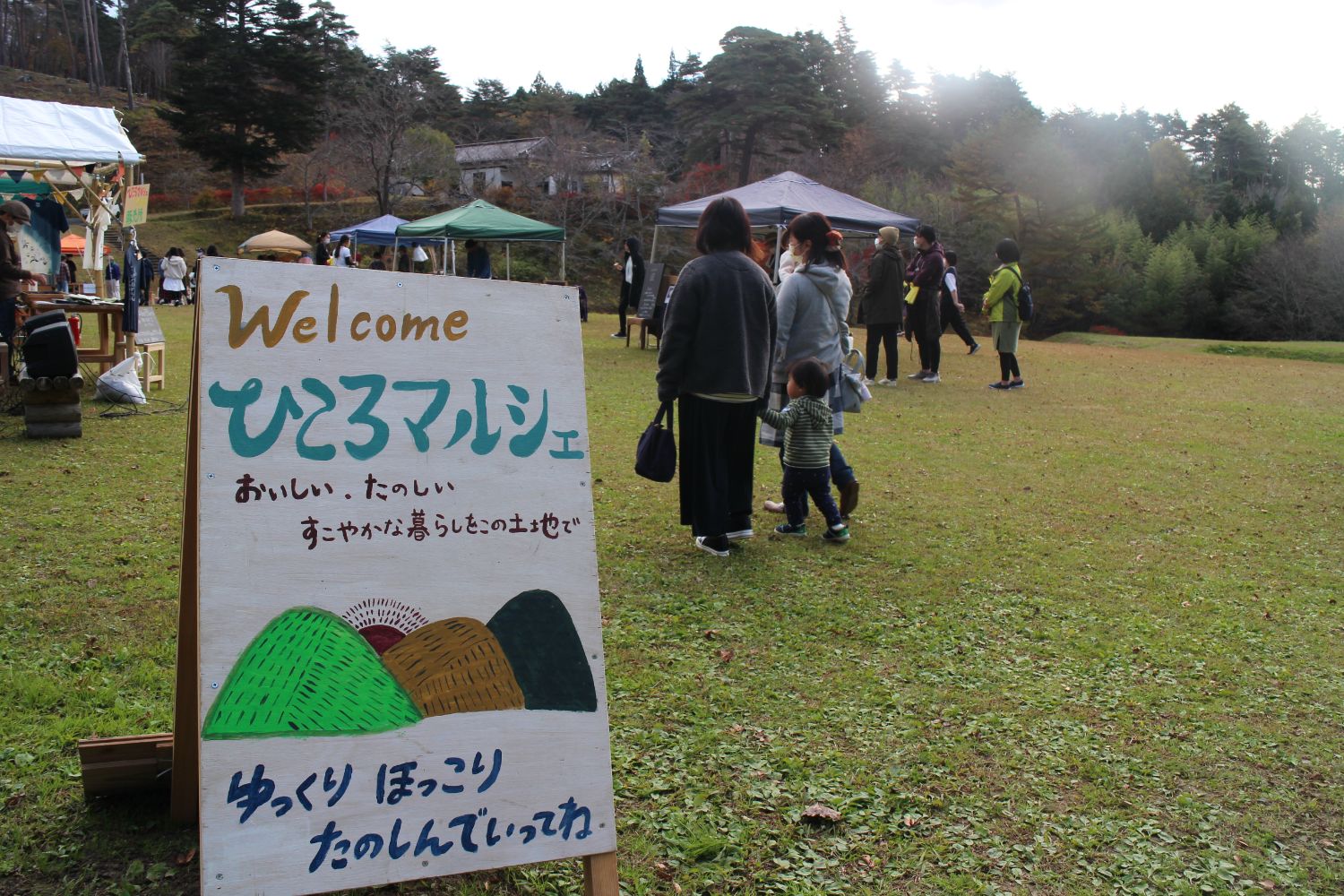 芝生の広場に「Welcome ひころマルシェ」と書かれた看板が置かれていて、その先に数軒のテントや人々が並び、秋の木々が背景に見えるイベント会場の風景写真