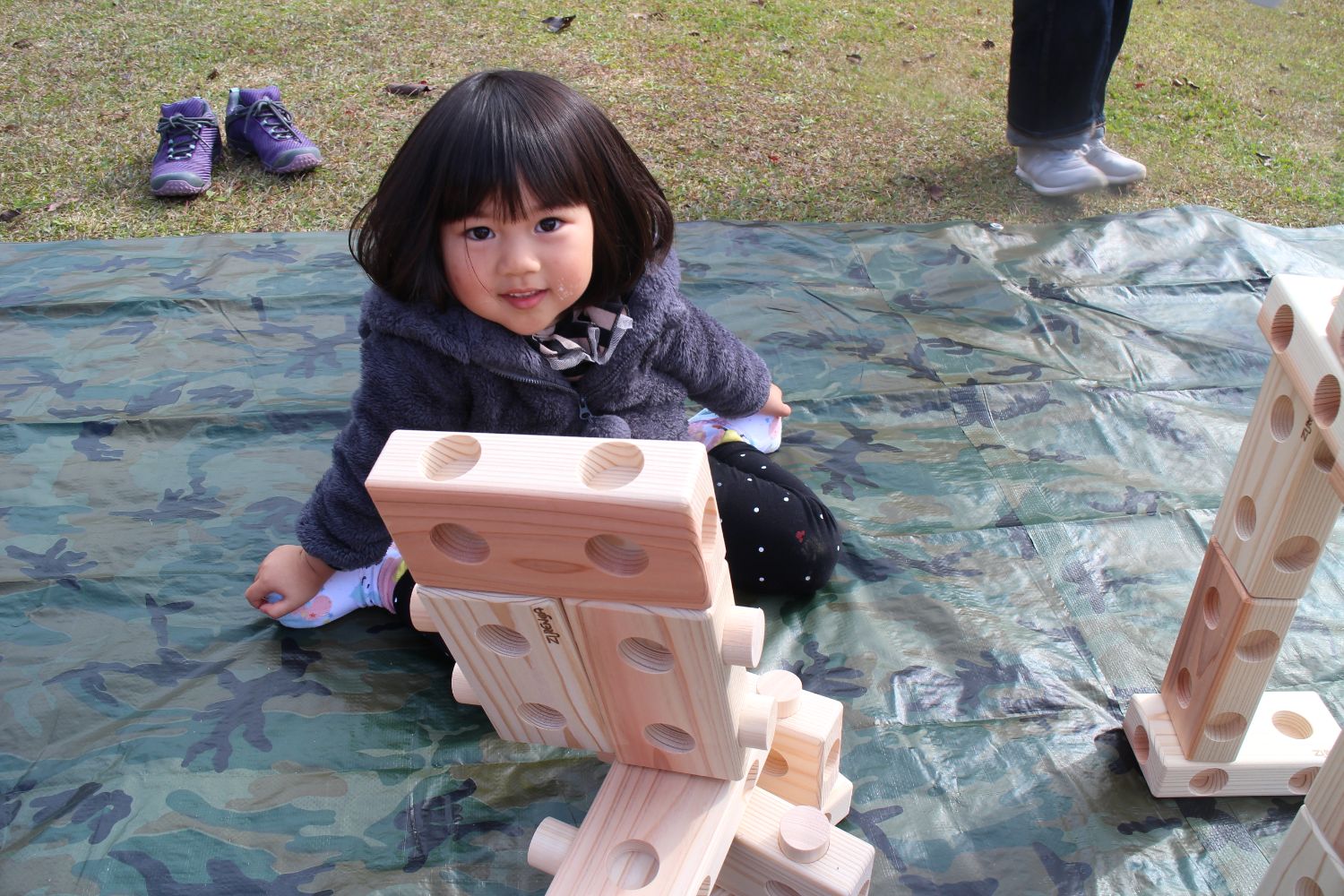 木製の積み木の傍に座る女の子がカメラ目線で微笑んでいる写真