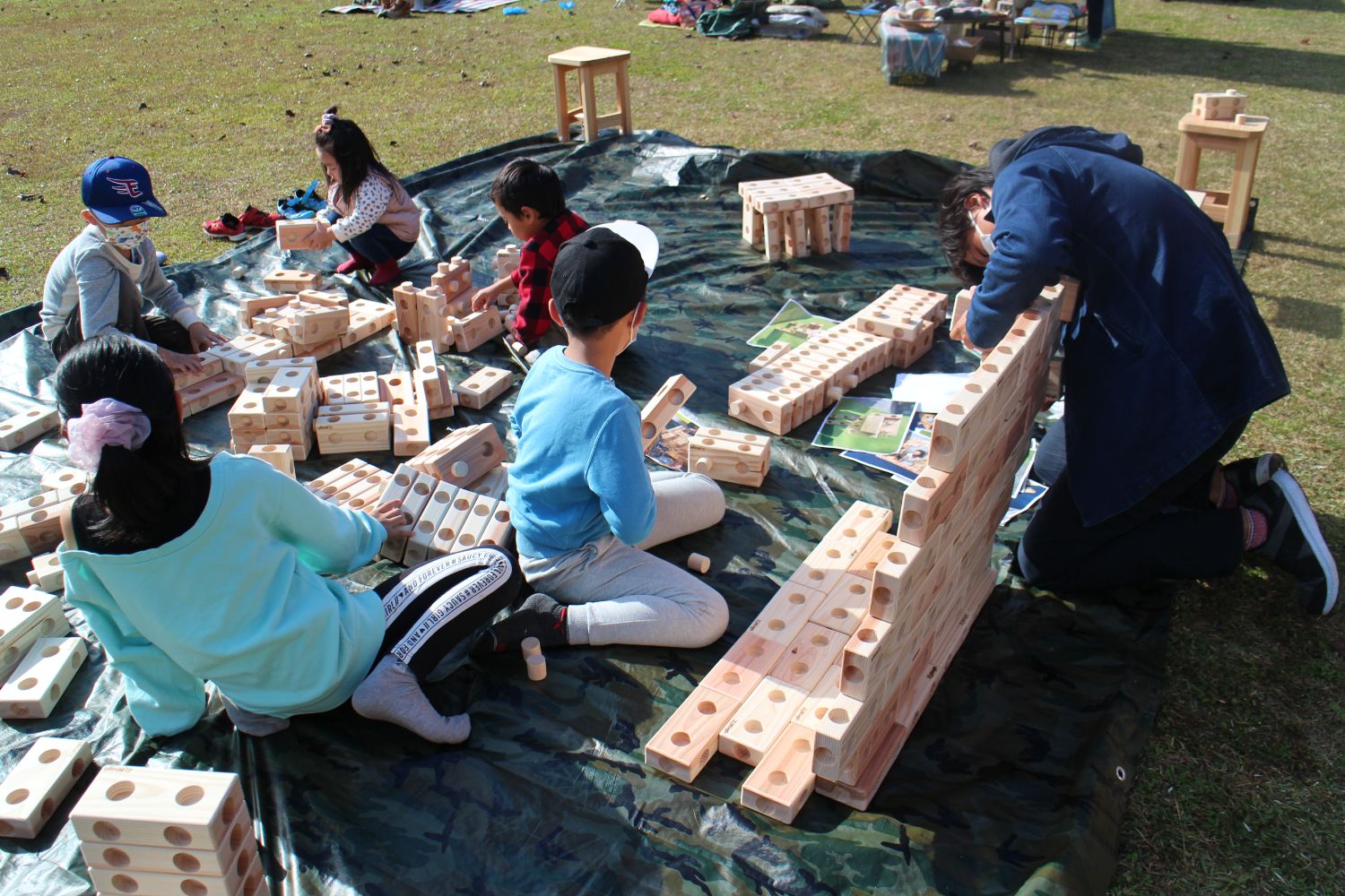 大きなシートの上で複数の子どもが木製の大きな積み木を使って遊んでおり、子どもと大人が一緒に積み木を組み立てている様子の写真