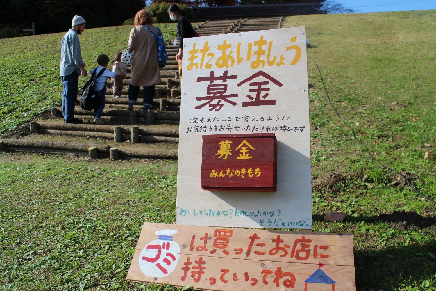 芝生の坂道に設置された木製の募金箱付き看板に「またあいましょう募金」と大きく書かれ、「次もまたここで会えるようにお気持ちをお寄せいただければ嬉しいです」とメッセージが添えられており、その下に「ゴミは買ったお店に持っていってね」と手書きの案内板が並べられていて、階段を登っていく来場者の姿が写っている写真