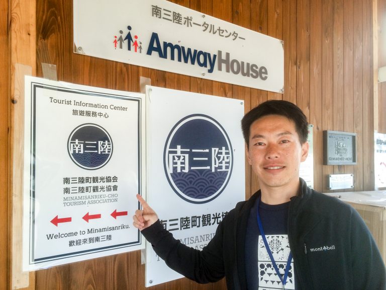 木の壁に設置された「南三陸ポータルセンター Amway House」の看板の下に「南三陸町観光協会」の看板があり、その左側に掲示された英語や中国語などが併記された観光協会の張り紙を指差し、笑顔で立っている男性の写真