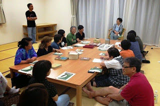 本が置かれた和室の座卓を囲んで座っている十数人の人たちが、奥の椅子に座っている高齢の女性の話を熱心に聞いている写真