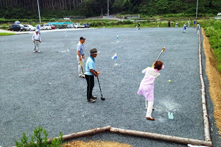 広場でピンク色の服を着た女性がクラブを使ってボールを打ち、それを他の数人の人達が見ているグラウンドゴルフを楽しんでいる様子の写真