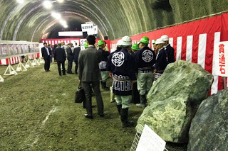 壁に紅白幕が飾られたトンネル内で、法被とヘルメットを着用した作業員たちが集まり、奥ではスーツ姿の来賓が立ち話をしている写真