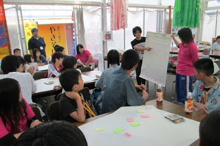 参加者の子どもたちがテーブルを囲み、MVCぶらんこ代表の方が前で大きな紙に書かれた内容を指し示しながら説明しているワークショップの写真