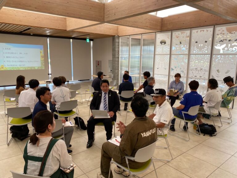 会議室で参加者たちが複数のグループに分かれて円になり、スーツ姿の人や作業服の人など多様な立場の人々が真剣に話し合っている様子の写真