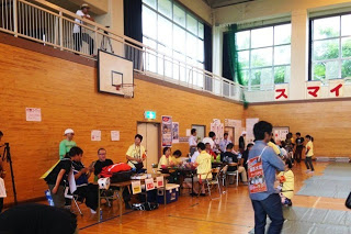 体育館の壁際にテーブルが並べられ、黄色いTシャツを着たスタッフたちが座って対応している様子の写真