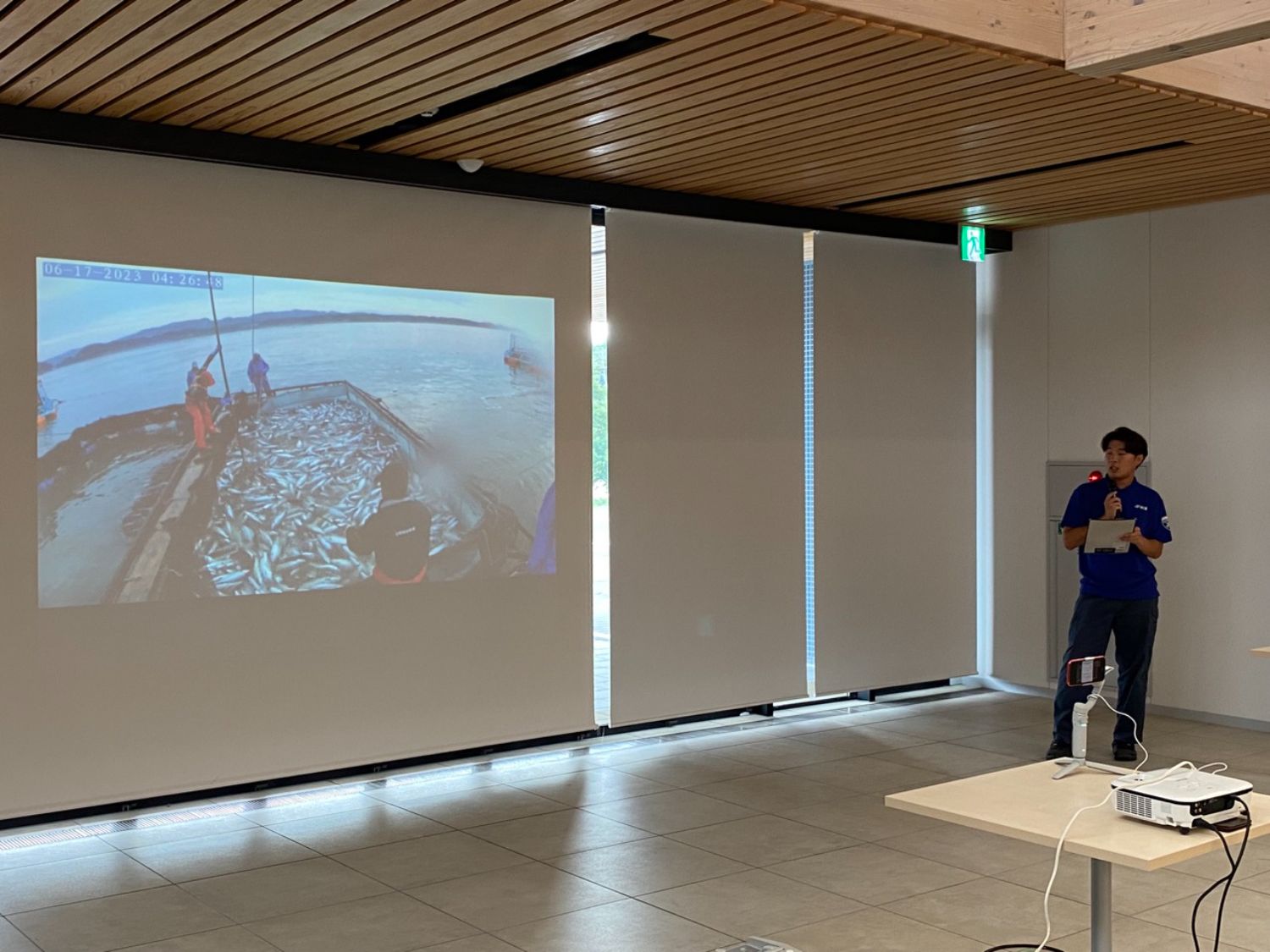 男性が室内で魚の漁獲風景を映したスライドを示しながらプレゼンテーションしている写真