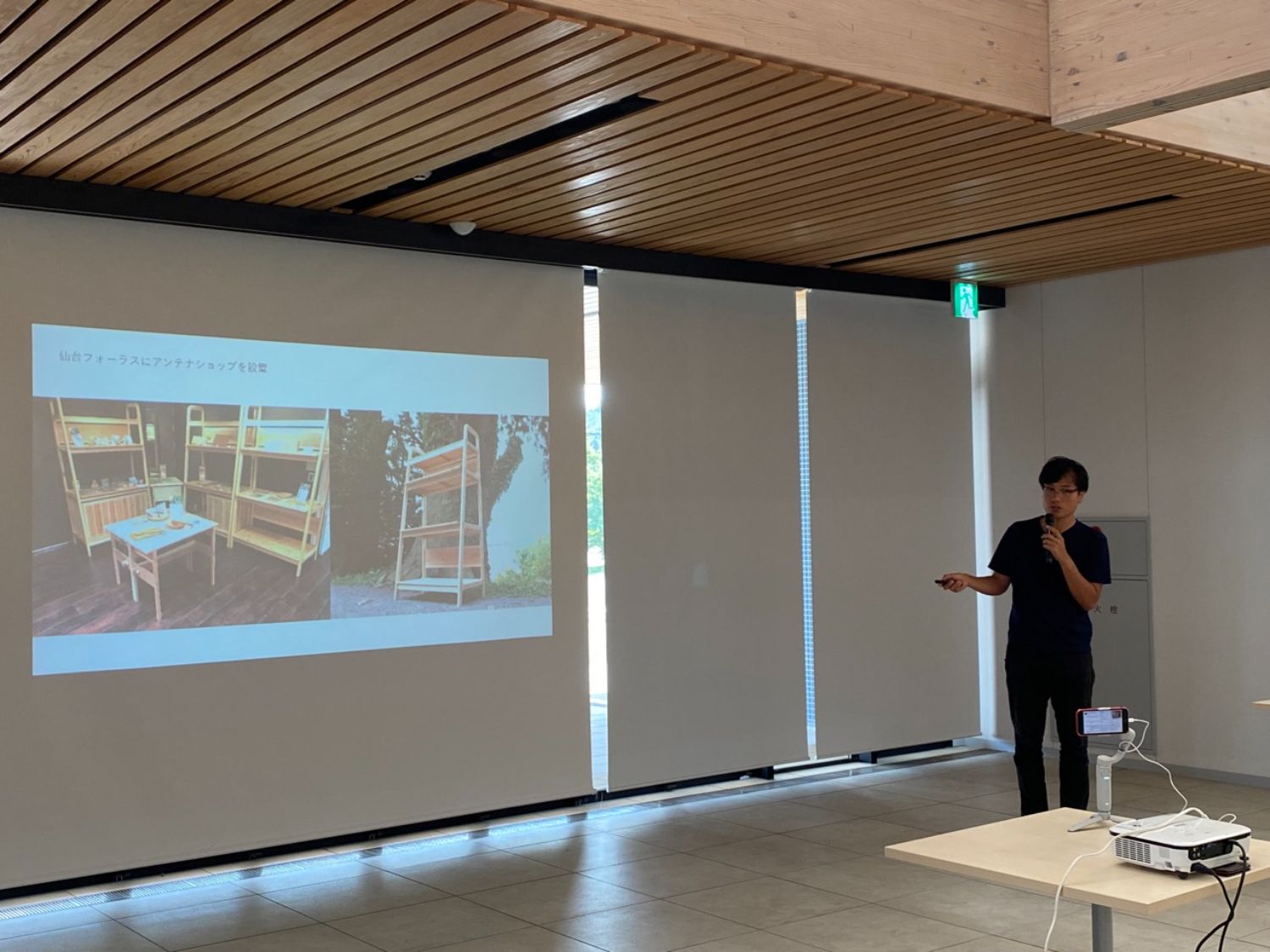 男性が黒い服を着て、マイクを持ちながらスライドを指し示して説明している写真