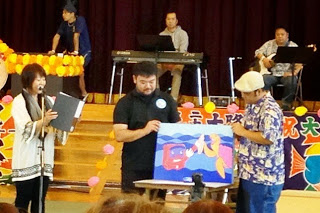 ステージ上で3人がマイクを使って話しながら、鮮やかな色のイラストパネルを披露している様子の写真