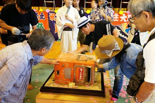 舞台上で人々が古い赤いポストを囲んで懐かしそうに見つめている様子の写真