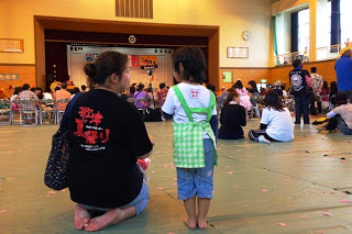 ステージを正面に見ながら親子が床に座って祭りを楽しんでいる様子の後ろ姿の写真