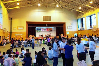 体育館の中央に人々が集まり、前方のステージでは音楽バンドが演奏している様子を写した夏祭りの室内会場の写真