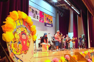 「歌津夏まつり」と書かれた横断幕や、カラフルな装飾がされたステージ上で、バンド演奏が行われている様子の写真