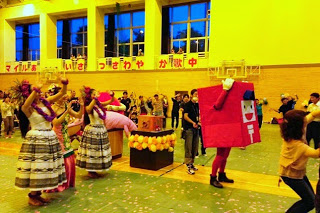 体育館の床の上でフラダンスを踊る女性たちと、赤いポストの着ぐるみが並んでいる様子の写真