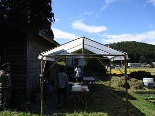 緑に囲まれた畑のそばの木造小屋の前に簡易的なテントが設置され、人々が中で準備作業をしている写真